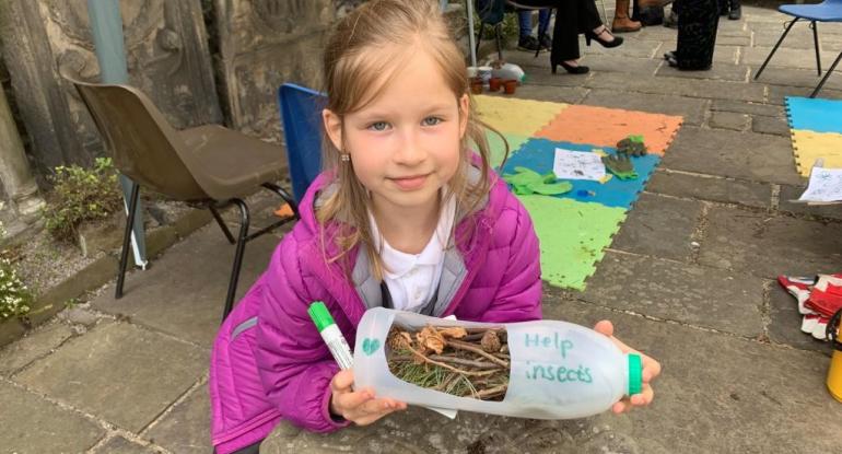 A girl in a purple coat holding a plastic milk bottle that has been turned into a bug hotel with 'save the insects' written on it