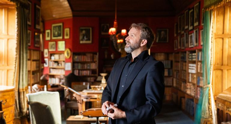 A magician holding a pack of cards standing in the library at Lauriston. The walls are covered in books and pictures.