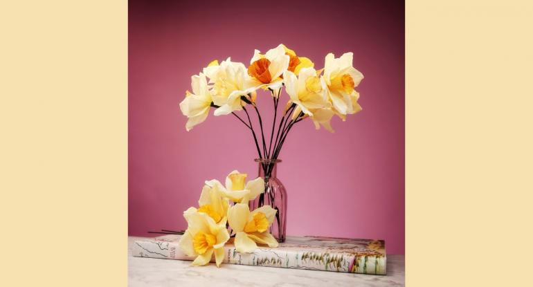 A glass jar of paper daffodils, with loose ones lying beside the jar on a  book