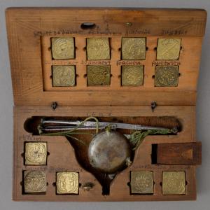 Close up of a wooden box with embossed brass square coin weights and a small weighing scale with metal pans