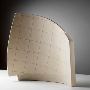 A modern, abstract sculpture featuring a curved beige surface divided into a grid pattern, standing against a simple grey background.